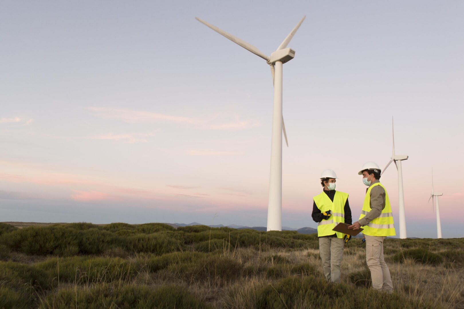 a couple of men wearing safety vests and helmets standing in a field with a wind turbine